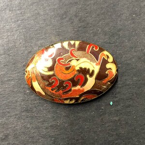 May include: An oval-shaped brooch with a gold-toned metal base and a colorful enamel design. The design features swirls and curves in shades of brown, orange, red, and white.