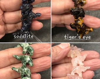 May include: Four strands of gemstone beads, each featuring a different color and type of stone. The beads are shaped like elephants with raised trunks and ears. The stones are sodalite, tiger's eye, tree agate, and rose quartz.