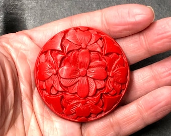 Può includere: Un bottone rotondo di colore rosso, intagliato con un motivo floreale. Il bottone è realizzato in un materiale duro e liscio e ha una finitura lucida.