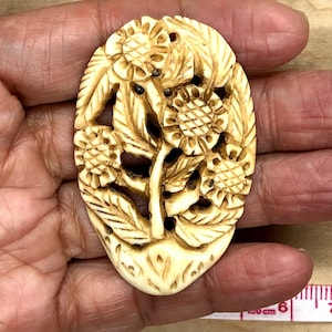 May include: A carved oval pendant made of bone with a detailed floral design. The pendant features a central stem with leaves and multiple flowers with intricate carvings.