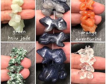 May include: A collection of six strands of gemstone beads, each strand featuring a different type of stone. The strands are green hsiu jade, orange aventurine, tree agate, sodalite, and clear quartz. Each strand is strung with a white thread and features a unique, carved design.