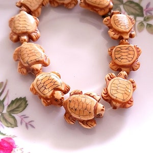 May include: A string of beige turtle-shaped beads. The beads are carved with detailed shell patterns and have a slightly glossy finish.