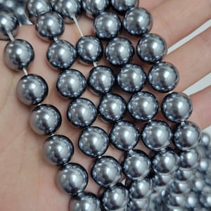 May include: A strand of round, metallic dark gray beads with a smooth, reflective surface. The beads are uniform in size and shape, strung together on a thin, clear filament, creating a continuous line of polished, spherical elements.