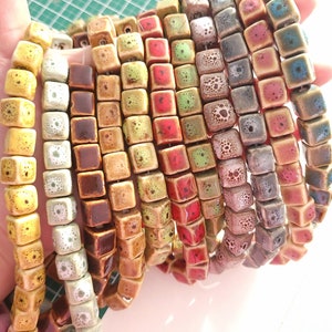 May include: A collection of ceramic square beads in various colors, including yellow, brown, green, red, and blue. The beads have a textured surface and are arranged in a stack.