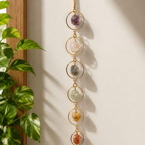 May include: A vertical wall hanging featuring six gold-toned metal rings connected by a gold chain. Each ring contains a raw, wire-wrapped gemstone in varying colors including purple, pink, blue, light green, yellow, and two shades of orange-red.