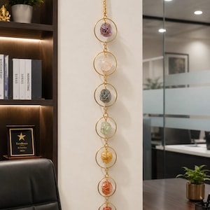 May include: A vertical wall hanging featuring seven gold-toned metal rings connected by small gold chains. Each ring contains a single raw, wire-wrapped gemstone in varying colors including purple, white, blue, green, yellow, orange, and red.