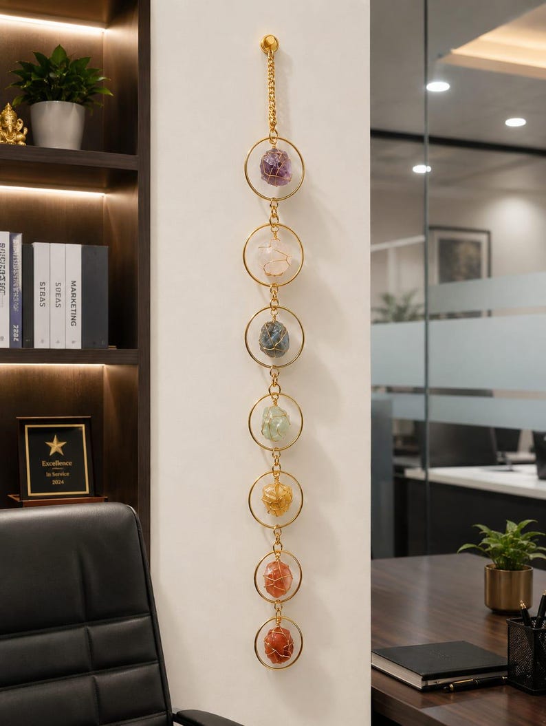 May include: A vertical wall hanging featuring seven gold-toned metal rings connected by small gold chains. Each ring contains a single raw, wire-wrapped gemstone in varying colors including purple, white, blue, green, yellow, orange, and red.