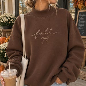 May include: A dark brown long-sleeve sweatshirt with a mock neck collar. The front features the word fall embroidered in a light-colored dashed script font, positioned above a simple embroidered line-art bow graphic in the same style.