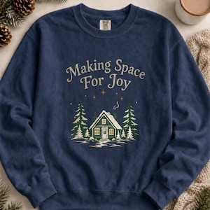 May include: A navy blue crewneck sweatshirt featuring the text "Making Space For Joy" in a light tan font above a graphic of a snow-covered cabin surrounded by pine trees, with small gold stars scattered in the night sky above the cabin.