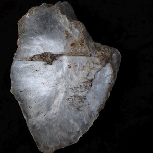 May include: A translucent, irregular slab of white quartz mineral with a polished, smooth surface. The stone features visible internal fractures, cloudy white inclusions, and small patches of brown mineral deposits across its jagged edges.