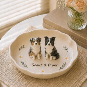 Puede incluir: Un plato redondo de cerámica color crema con bordes festoneados que presenta dos figuras de perro, una marrón y blanca y otra negra y blanca. El plato está decorado con margaritas blancas y hojas verdes, con el texto Scout & Piper.