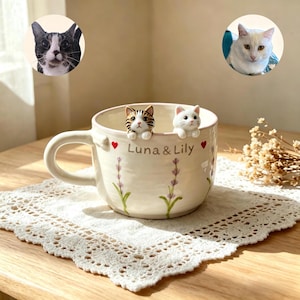 Puede incluir: Una taza de cerámica color crema con dos pequeñas figuras de gato en el borde, una atigrada marrón y otra blanca. La taza está decorada con ramas de lavanda pintadas, corazones rojos y el texto "Luna & Lily" impreso en la parte frontal.