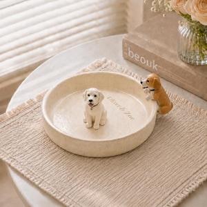 May include: A round, cream-colored ceramic bowl with a speckled texture. A small white dog figurine sits in the center, and a brown and white dog figurine clings to the rim. The interior base features the engraved text Oliver & Zoe.