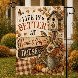 Puede incluir: Una bandera de jardín rectangular con fondo de veta de madera y el texto "LIFE IS BETTER AT NANA & PAPA'S HOUSE". El diseño incluye una pajarera, un pájaro, una mariposa, girasoles, una regadera de metal, calabazas y hojas de otoño.