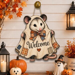 May include: A decorative wall hanging shaped like an opossum wearing a cream-coloured blanket with a plaid bow tie. The blanket features the word Welcome in black script, a small orange heart, and various plaid patches in orange, blue, and brown.