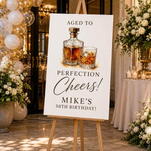 May include: A white rectangular sign on a wooden easel featuring a watercolor illustration of a glass decanter and a glass of amber liquid. The text reads: AGED TO PERFECTION Cheers! MIKE'S 50TH BIRTHDAY! The sign has a clean, professional layout.