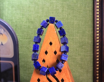 Könnte beinhalten: Ein Armband bestehend aus abwechselnden tiefblauen, würfelförmigen Lapislazuli-Perlen und kleinen, runden, dunkelblauen Zwischenperlen. Das Armband ist oval angeordnet und zeigt die natürliche, gesprenkelte Textur der Steinwürfel sowie die glatte Oberfläche der kleineren runden Perlen.
