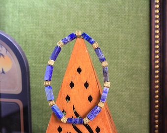 Könnte beinhalten: Ein Armband aus abwechselnd rechteckigen, blauen Lapislazuli-Steinen und kleinen, quadratischen, goldfarbenen Metall-Abstandshaltern mit einem komplizierten geprägten Muster. Die Steine haben ein tiefes Blau mit natürlichen grauen Einschlüssen.