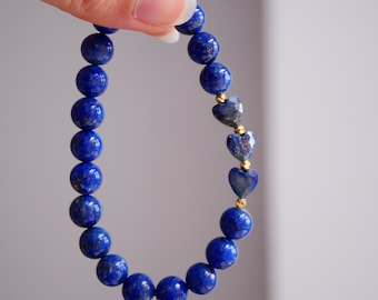 Könnte beinhalten: Ein Perlenarmband mit tiefblauen, kugelförmigen Lapislazuli-Perlen und drei facettierten, herzförmigen Lapislazuli-Perlen. Kleine, runde, goldfarbene Metall-Abstandshalter befinden sich zwischen den herzförmigen Steinen.