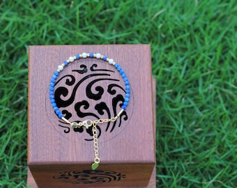 Könnte beinhalten: Ein zartes Armband mit kleinen, runden, leuchtend blauen Perlen, die mit winzigen weißen Perlen und metallisch goldfarbenen Akzenten durchsetzt sind. Das Schmuckstück verfügt über eine goldfarbene Verlängerungskette mit Karabinerverschluss und einen kleinen blattförmigen Anhänger.