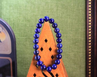 Könnte beinhalten: Ein kreisförmiges Armband aus glatten, runden, tiefblauen Lapislazuli-Perlen. Die Perlen sind von einheitlicher Größe und auf ein elastisches Band gefädelt, wodurch eine durchgehende Schlaufe entsteht, die auf einem dreieckigen Holzständer präsentiert wird.
