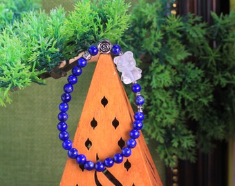 Könnte beinhalten: Ein Perlenarmband mit runden, tiefblauen Lapislazuli-Steinen, einem silberfarbenen Metallanhänger in Rosenform und einem klaren, durchscheinenden Anhänger in Bärenform. Die Perlen sind mit kleinen silberfarbenen Metallzwischenstücken verbunden.