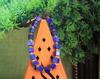 Könnte beinhalten: Ein Armband aus tiefblauen, würfelförmigen Lapislazuli-Steinen, die sich mit kleinen, metallisch goldfarbenen Zwischenperlen abwechseln. Das Armband ist oval angeordnet und zeigt die einheitliche Größe und Farbe der Steine.
