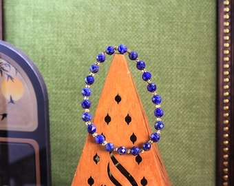 Könnte beinhalten: Ein Perlenarmband mit runden, facettierten, tiefblauen Lapislazuli-Steinen, die sich mit kleinen, polierten goldfarbenen Metall-Abstandshaltern abwechseln. Das Armband ist kreisförmig angeordnet und auf einem Holzständer präsentiert.