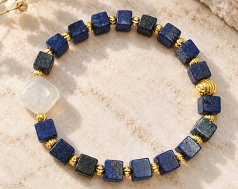 Könnte beinhalten: Ein Perlenarmband mit dunkelblauen, quadratischen Lapis-Lazuli-Perlen, die sich mit kleinen goldfarbenen Metallkugeln abwechseln. Eine größere, glatte, durchscheinend weiße quadratische Perle befindet sich auf der linken Seite des elastischen Bandes.