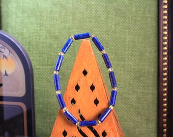Könnte beinhalten: Ein Armband mit zylindrischen, tiefblauen Lapislazuli-Perlen, die sich mit kleinen, runden, metallisch goldfarbenen Zwischenperlen abwechseln. Die Perlen sind in einem kreisförmigen Muster aufgereiht und bilden eine durchgehende Schleife.