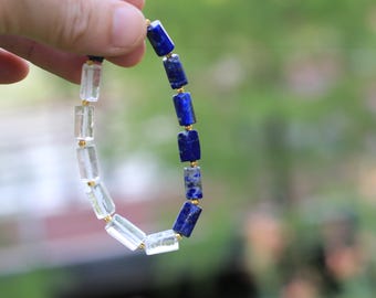 Könnte beinhalten: Ein Armband mit abwechselnden zylindrischen Perlen aus tiefblauem Lapislazuli und klarem, transparentem Quarz. Die Perlen sind in einem sich wiederholenden Muster angeordnet und durch kleine, goldfarbene Metallzwischenperlen auf einer Schnur getrennt.