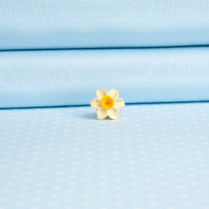 May include: A white ceramic flower with a yellow center, resembling a daffodil, is placed on a light blue fabric with white polka dots.