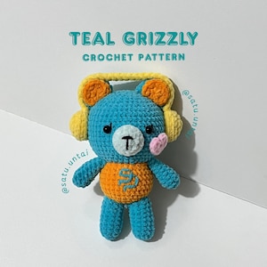 May include: A teal crochet grizzly bear plushie wearing yellow headphones, featuring an orange belly with a blue embroidered symbol, a white muzzle with a black nose and mouth, and a small pink heart-shaped cheek. Text reads TEAL GRIZZLY CROCHET PATTERN.