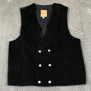 May include: A black double-breasted vest featuring a subtle white polka dot pattern. The garment has a V-neckline, six light-colored buttons arranged in two vertical rows, and a black interior lining with a rectangular brand label at the neck.