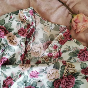 May include: A white satin robe with a floral print featuring pink and red roses. The robe has a V-neckline and a label that reads "Linda's Boutique".