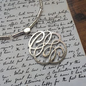 K&ouml;nnte beinhalten: Silberfarbene Halskette mit einem gro&szlig;en, durchbrochenen Anh&auml;nger mit einem verzierten, stilisierten Buchstaben-Design. Die Halskette hat eine Perlenkette mit klaren, facettierten Perlen, pr&auml;sentiert auf einem handgeschriebenen Papierhintergrund.