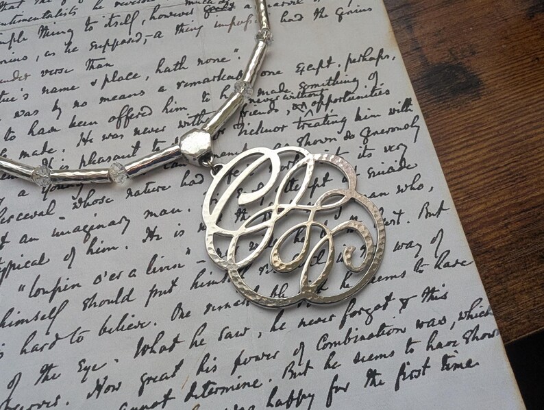 K&ouml;nnte beinhalten: Silberfarbene Halskette mit einem gro&szlig;en, durchbrochenen Anh&auml;nger mit einem verzierten, stilisierten Buchstaben-Design. Die Halskette hat eine Perlenkette mit klaren, facettierten Perlen, pr&auml;sentiert auf einem handgeschriebenen Papierhintergrund.