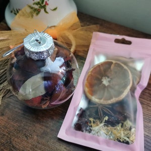 May include: A clear glass ornament filled with dried fruit, spices, and herbs. A pink pouch with a dried orange slice, star anise, and other spices is next to the ornament.