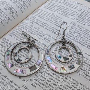 May include: A pair of silver earrings with a double circle design. The circles are made of white metal with a mother-of-pearl inlay. The earrings are hanging from a book with text about a knight named Triamond.