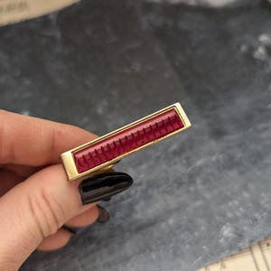 May include: A gold-toned barrette with a red, rectangular, textured glass insert.