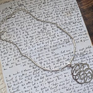 K&ouml;nnte beinhalten: Eine silberfarbene Halskette mit einem floralen Anh&auml;nger, die auf einem Vintage-Handschriften-Dokument liegt. Die Halskette hat eine Kette mit kleinen, zylindrischen Perlen und einen dekorativen, durchbrochenen Anh&auml;nger.