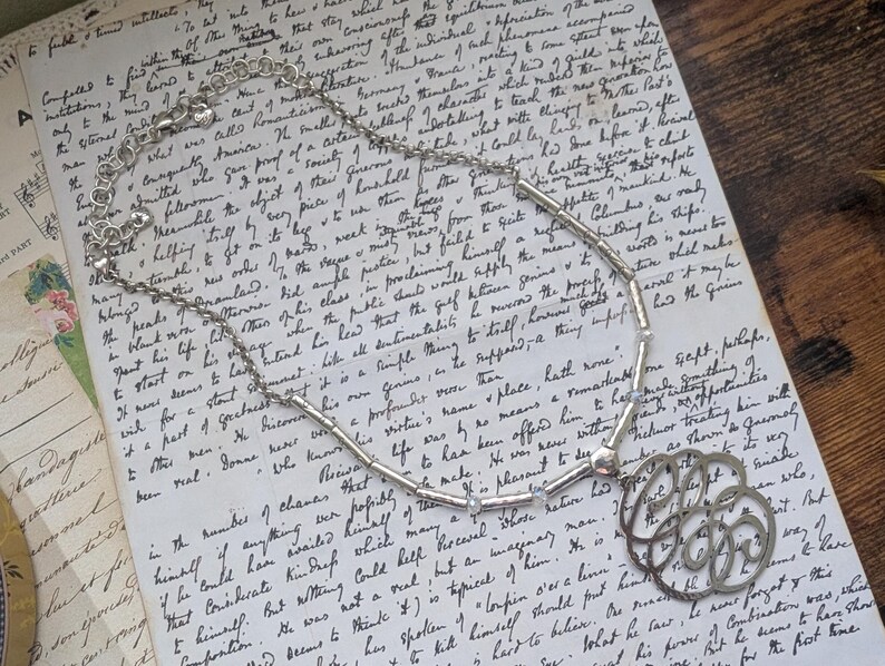 K&ouml;nnte beinhalten: Eine silberfarbene Halskette mit einem floralen Anh&auml;nger, die auf einem Vintage-Handschriften-Dokument liegt. Die Halskette hat eine Kette mit kleinen, zylindrischen Perlen und einen dekorativen, durchbrochenen Anh&auml;nger.