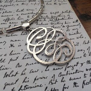 K&ouml;nnte beinhalten: Eine silberfarbene Halskette mit einem gro&szlig;en, verzierten Anh&auml;nger mit verschlungenen Initialen. Der Anh&auml;nger h&auml;ngt an einer Perlenkette und liegt auf einem handgeschriebenen Vintage-Dokument. Die Halskette hat eine geh&auml;mmerten Textur und ein gl&auml;nzendes Finish.