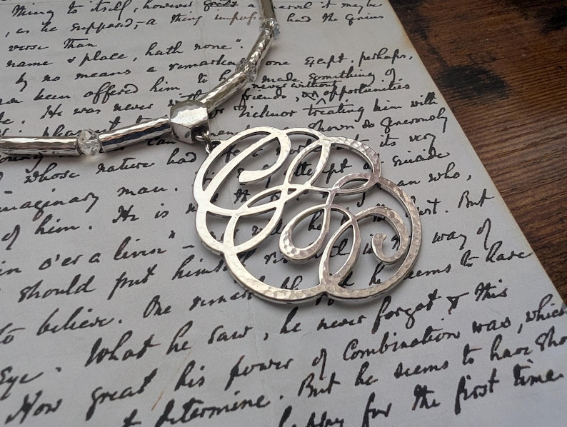 K&ouml;nnte beinhalten: Eine silberfarbene Halskette mit einem gro&szlig;en, verzierten Anh&auml;nger mit verschlungenen Initialen. Der Anh&auml;nger h&auml;ngt an einer Perlenkette und liegt auf einem handgeschriebenen Vintage-Dokument. Die Halskette hat eine geh&auml;mmerten Textur und ein gl&auml;nzendes Finish.
