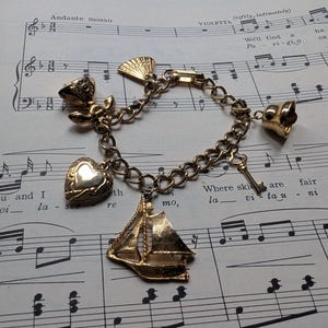 May include: A gold-tone charm bracelet featuring five distinct charms: a sailboat, a heart-shaped locket, a small bell, a fan, and a key. The bracelet is constructed with a classic link chain design, showcasing a polished metallic finish throughout.