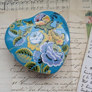 K&ouml;nnte beinhalten: Eine herzf&ouml;rmige Schmuckschatulle mit floralem Design in Blau, Gelb und Wei&szlig;. Die Schatulle hat einen t&uuml;rkisfarbenen Hintergrund und das Wort "Brighton" ist auf dem Deckel zu sehen. Sie liegt auf einem Vintage-Papierhintergrund.