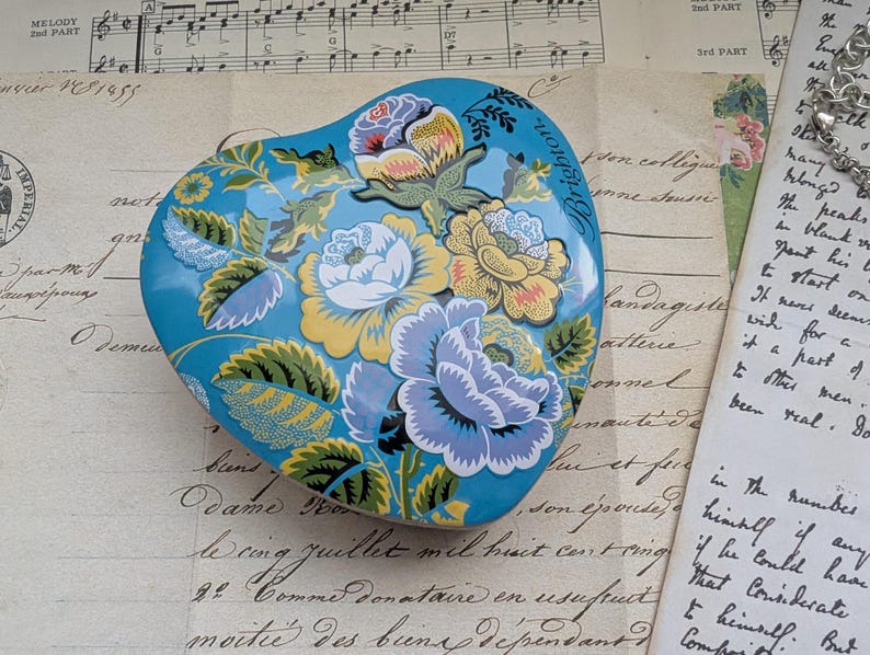 K&ouml;nnte beinhalten: Eine herzf&ouml;rmige Schmuckschatulle mit floralem Design in Blau, Gelb und Wei&szlig;. Die Schatulle hat einen t&uuml;rkisfarbenen Hintergrund und das Wort "Brighton" ist auf dem Deckel zu sehen. Sie liegt auf einem Vintage-Papierhintergrund.