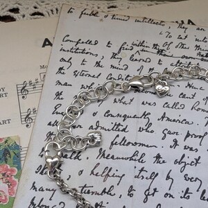 K&ouml;nnte beinhalten: Ein silberfarbenes Armband mit einem herzf&ouml;rmigen Anh&auml;nger und einem Karabinerverschluss. Das Armband liegt auf einem Vintage-Papierhintergrund mit handschriftlichem Text und Noten. Die Kette des Armbands besteht aus runden Gliedern.