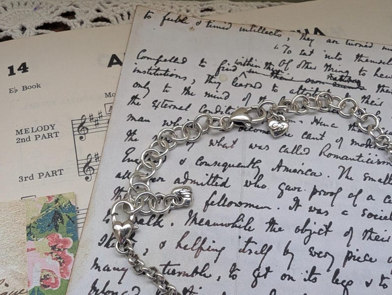 K&ouml;nnte beinhalten: Ein silberfarbenes Armband mit einem herzf&ouml;rmigen Anh&auml;nger und einem Karabinerverschluss. Das Armband liegt auf einem Vintage-Papierhintergrund mit handschriftlichem Text und Noten. Die Kette des Armbands besteht aus runden Gliedern.