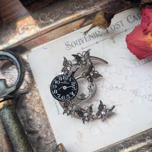 May include: A silver crescent moon brooch with a clock face and a detailed design of butterflies and stars. The brooch is on a vintage postcard with the text "Souvenir Post Card".